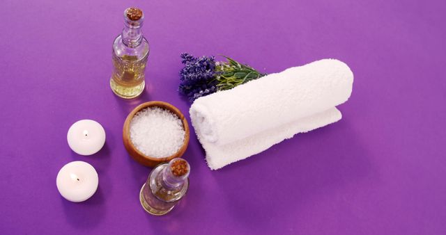 This image depicts a serene aromatherapy setup featuring essential oils, bath salts, a rolled white towel, lavender sprigs, and lit candles on a purple background. Perfect for promoting spa services, wellness products, relaxation techniques, or self-care routines.