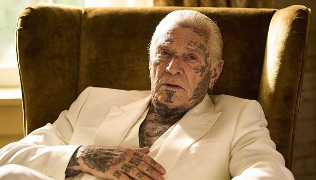 Tattooed senior man reclining in cream suit on velvet wingback chair closeup portrait