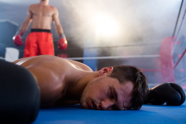 This image captures the dramatic moment of a boxer lying unconscious in the ring after a knockout. The scene is intense, with another athlete standing in the background, highlighting the competitive nature of the sport. Ideal for use in articles or advertisements related to boxing, sports training, fitness motivation, or sportsmanship. It can also be used in promotional materials for gyms, boxing clubs, or sports events.