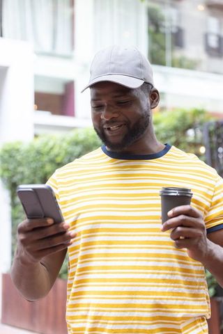 African american man smiling while checking smartphone and holding takeaway coffee