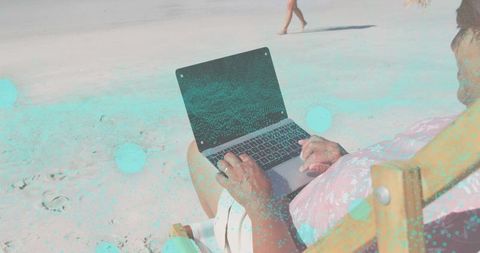 Senior woman typing on laptop on beach chair with turquoise overlay, sunlit sand, copy space