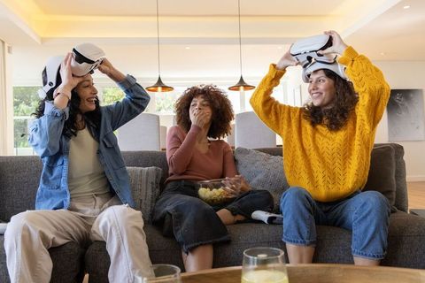 Diverse women trying vr headsets on sofa laughing and sharing snacks during game night