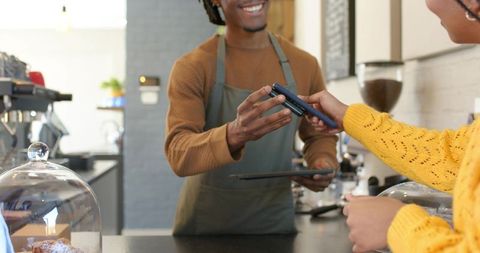 Smiling african american barista taking contactless smartphone payment at cafe counter