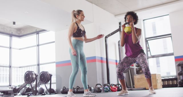 Image of diverse female fitness trainer instructing woman doing squats with kettlebell weight at gym. Exercise, fitness and healthy lifestyle.