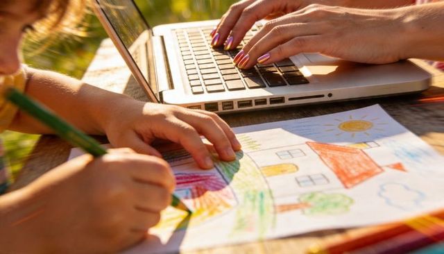 Parent working remotely while child coloring on wooden table in garden sunlight