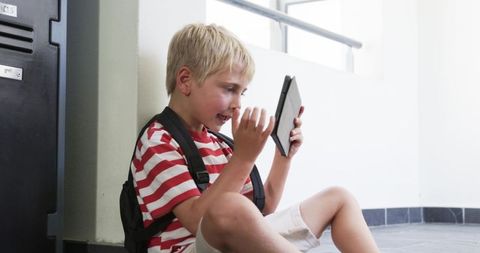 Young student using tablet while sitting in school hallway with backpack and lockers