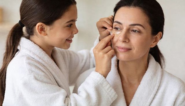 Daughter applying under-eye gel patch on mother during morning skincare routine