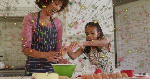 Joyful mother and daughter baking together cracking eggs into green mixing bowl on kitchen island