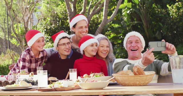 Multi-generational family wearing Santa hats during outdoor Christmas celebration, happily taking selfie together. Can be used for holiday greeting cards, festive advertisements, or articles on family traditions and Christmas celebrations.