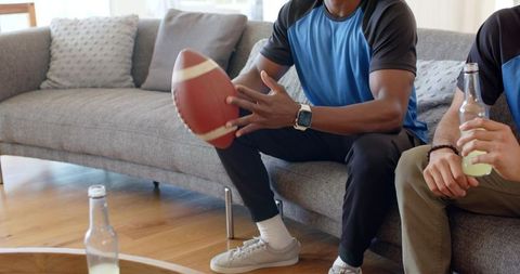 Diverse friends holding football and drinks on gray sofa watching game together