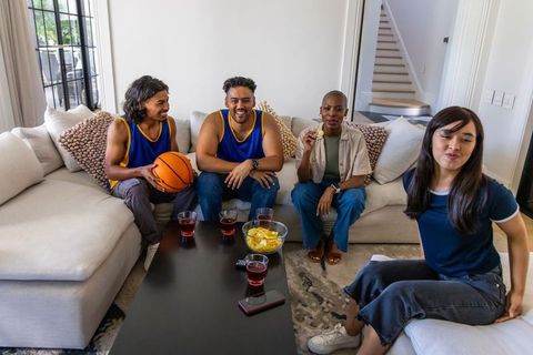 Friends watching basketball game at home cheering, holding ball, snacking on sofa