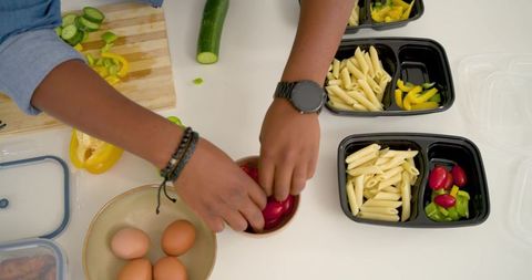 Hands preparing meal prep containers with pasta, cherry tomatoes, sliced peppers and eggs