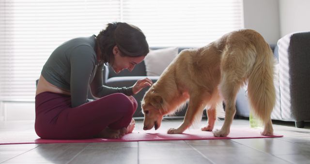 A woman is practicing yoga on a mat in her living room while interacting with her dog. This photo showcases the bond between pets and their owners and the integration of wellness and fitness into home life. It is suitable for use in lifestyle blogs, pet care articles, home fitness tutorials, and promotions for yoga or wellness products.