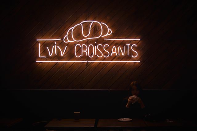 Neon sign reading 'Lviv Croissants' glowing on dark wooden wall in a dimly lit café. Quiet atmosphere highlights the sign as a focal point in the room. Suitable for marketing cafes, restaurants, urban dining experiences, and cozy indoor settings. The dim lighting adds a warm, intimate feel ideal for promotional material emphasizing a modern, relaxed ambiance.