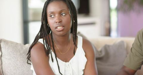 African woman sitting on sofa looking away wearing gold necklace, thoughtful home moment