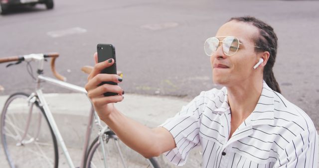 Front view close up of a biracial man with long dreadlocks out and about in the city on a sunny day, sitting in the street, wearing headphones, using a smartphone, waving his hand, with his bicycle standing next to him in slow motion.