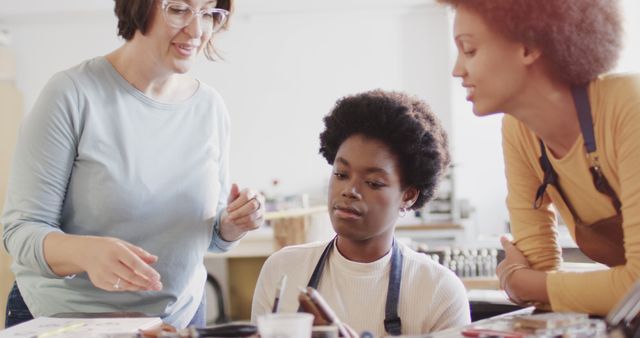 Teacher guiding female student on art project using painting supplies in a classroom. Woman in glasses and casual outfit helping young female student, another young woman observing. Perfect for educational, mentoring, or school-related themes.