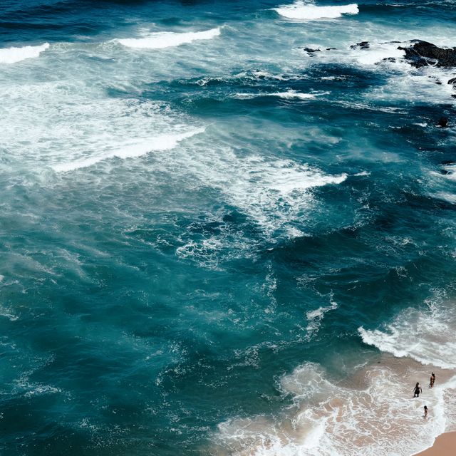 Aerial view of the turquoise ocean waves crashing onto a sandy beach with a few people enjoying the shore. This scene can be used for travel-related materials, promoting beach destinations, or illustrating concepts of relaxation and nature. Ideal for brochures, websites, and social media content.
