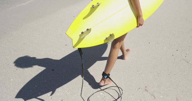 Person carrying bright yellow surfboard on sandy beach, ready to hit the waves. Great for websites or articles about beach activities, surf culture, summer sports, and coastal vacations.