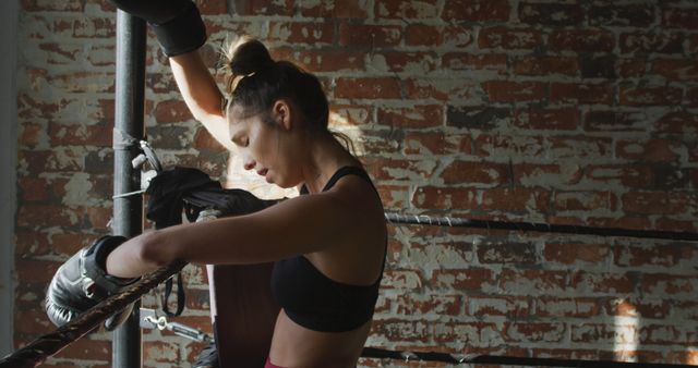 Female boxer in black sportswear leaning on ropes of a boxing ring, with exposed brick wall in background. This image can be used for fitness motivation, athletic training programs, sports advertisements, and health and wellness campaigns, emphasizing determination and hard work.