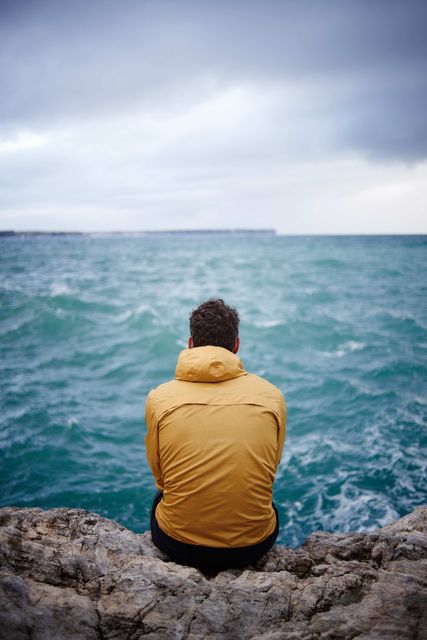 Person in yellow jacket sitting on a rocky shore, gazing at the sea. Cloudy sky and choppy waves. Ideal for themes of solitude, contemplation, travel, adventure, and nature scenes. Could be used in blogs, travel websites, mental health articles, or inspirational content.