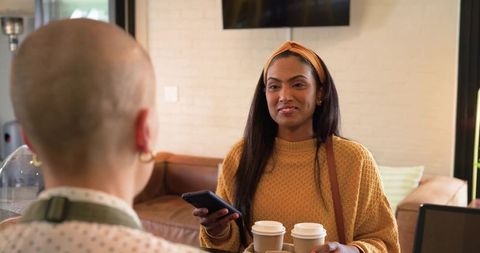 Indian woman ordering coffee at cafe counter holding phone and two takeaway cups