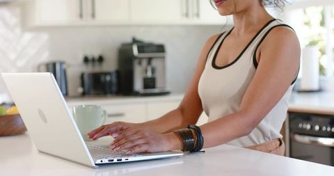 Indian woman typing on laptop at bright modern white marble kitchen island, remote work