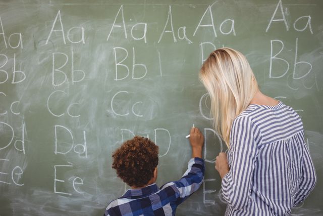 Teacher assisting schoolboy in writing alphabet on chalkboard at school