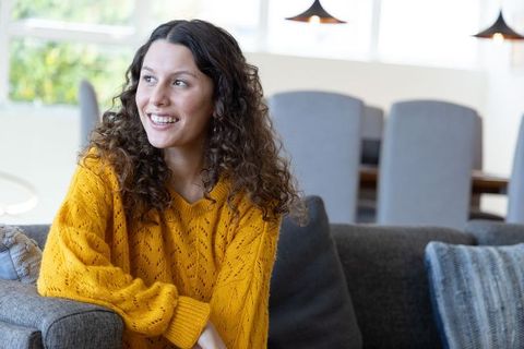 Young woman smiling on sofa wearing mustard sweater, relaxing in bright modern living room