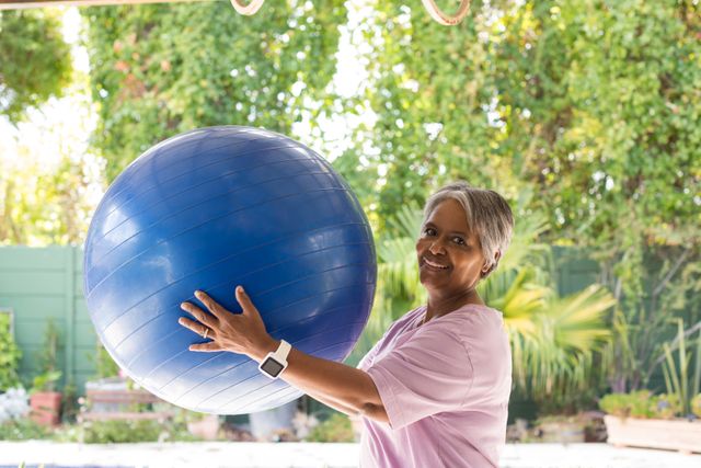 Senior woman holding a fitness ball in a garden, smiling and looking at the camera. Ideal for promoting healthy lifestyles, fitness programs for seniors, outdoor exercise routines, and wellness campaigns. The lush greenery in the background adds a refreshing and natural element to the scene.