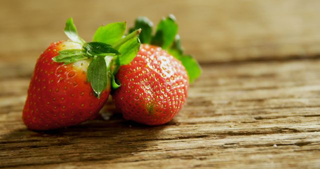 Closeup view of two ripe strawberries with green leaves placed on rustic wooden table. The natural setting adds a rustic feel, making the imagery perfect for promoting organic produce, healthy eating habits, farm-to-table concepts, recipe blogs, and summer fruit promotions.