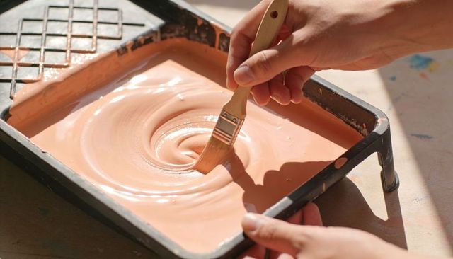 Stirring peach paint in tray with flat brush and hands creating glossy swirl in daylight