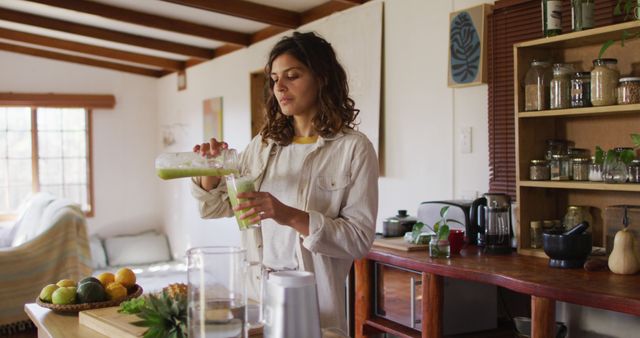 This depicts a young woman pouring a green smoothie into a glass in a cozy home kitchen. Ideal for promoting healthy lifestyle articles, morning routines, nutrition blogs, wellness campaigns, and home interior websites. Suggests a peaceful, health-conscious start to the day.