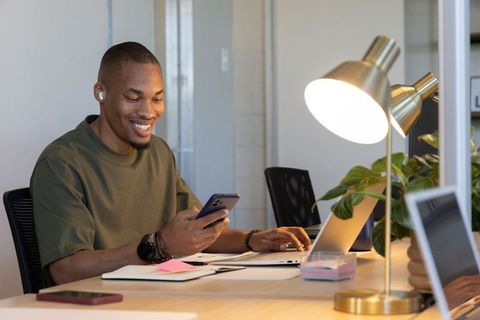 Smiling african american man working remotely checking smartphone at modern office desk