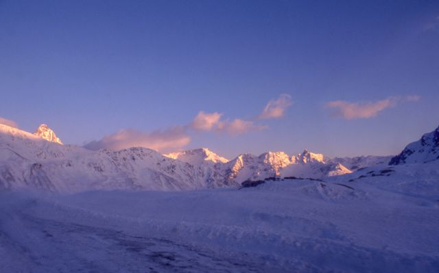 Snow-covered mountain range illuminated by the warm tones of the setting sun creating a vivid interplay with the purple sky. Suitable for promotional posters, nature magazines, and travel blogs, it can be used to convey tranquility, scenic beauty, and the serene coldness of winter landscapes.
