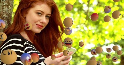 Redhead Woman Engaging with Smartphone Surrounded by Emojis in Park