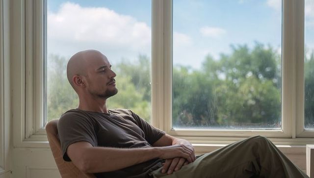 Relaxing bald man sitting in armchair by sunlit large window with leafy view, contemplative