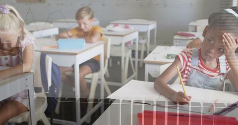Distracted elementary student writing while classmates concentrating in bright classroom