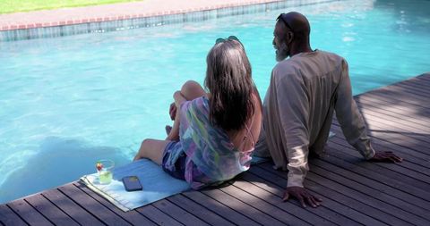 Multiracial mature couple sitting on backyard pool deck relaxing with drink and smartphone