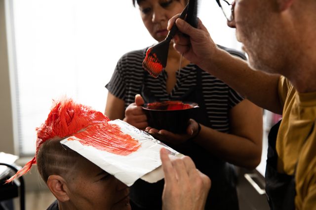 Hairdressers applying red dye to a client's hair in a salon. Useful for illustrating professional hair care services, beauty treatments, and teamwork in a salon setting. Ideal for beauty industry promotions, hair care product advertisements, and salon service brochures.
