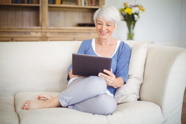 Senior woman sitting on a comfortable sofa in her living room, using a digital tablet. She is smiling and appears relaxed, enjoying her time browsing or reading. This image can be used for promoting technology use among seniors, lifestyle blogs, retirement planning, or home comfort products.
