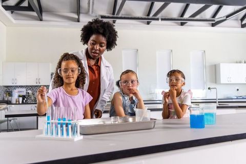 Female teacher guiding diverse girls during hands-on chemistry experiment with blue solution