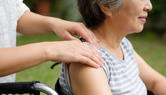Asian senior receiving gentle shoulder massage from caregiver in wheelchair outdoors