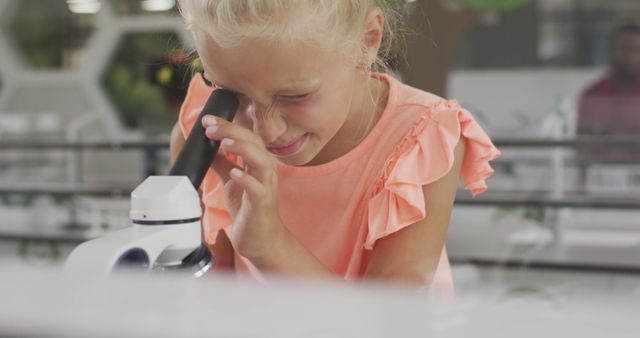 Young blonde girl is diligently examining a specimen using a microscope in a well-lit laboratory environment. This image is ideal for educational content, science-themed articles, or resources on children's STEM learning. It could also be used to promote school science programs, child development in scientific fields, or educational materials focused on biology and research.