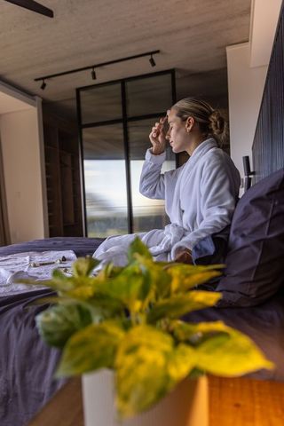 Woman in bathrobe sitting on bed massaging temple relaxing morning self care wellness moment