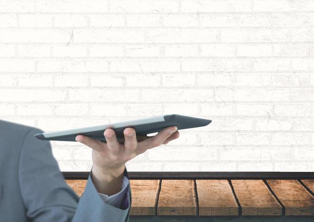 Businessman holding a tablet with a modern minimalist background consisting of a white brick wall and a wooden desk. This can be used to represent corporate, tech, and digital work themes, innovation in the workplace, or professional settings in presentations, advertisements, and websites.
