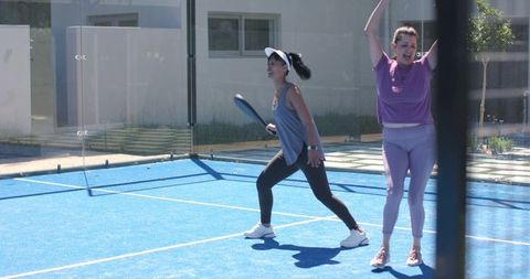 Women playing paddle tennis on bright blue court lunging, swinging, and celebrating point