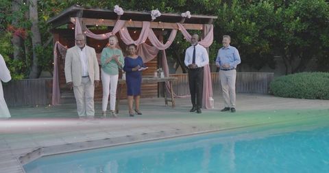 Mature adults celebrating poolside at pergola with pink drapery and tiered cake at dusk