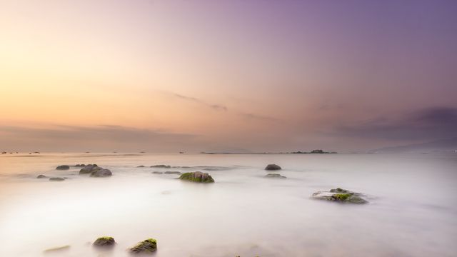 Gentle sunrise over tranquil coastal waters, creating a dreamy atmosphere with scattered rocks and misty water. Ideal for travel websites, nature blogs, inspirational posters, or serene desktop wallpapers.