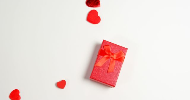 A red gift box with a bow is surrounded by small red hearts on a white background, with copy space. It suggests a romantic occasion such as Valentine's Day or an anniversary, emphasizing the concept of giving and love.
